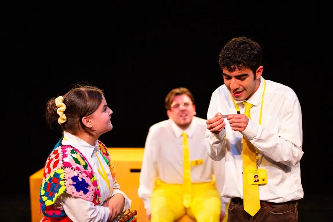Two men in white shirts and yellow ties interact with a young girl wearing a colourful cardigan on stage during a theatrical performance.
