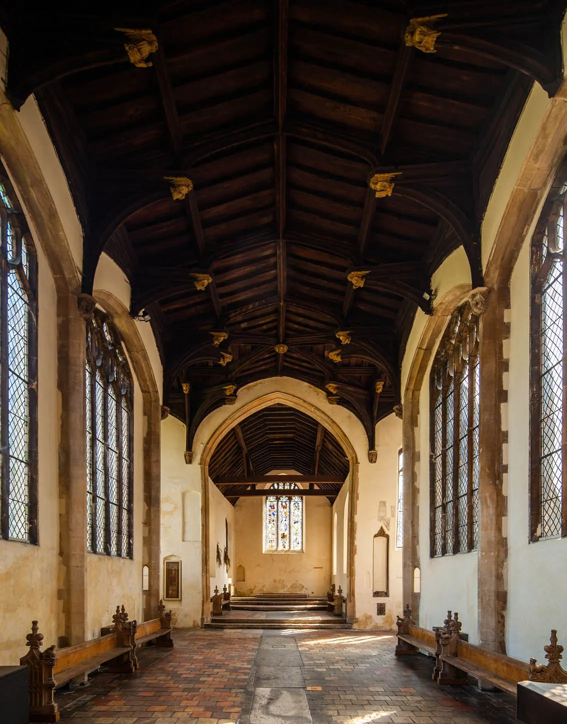 Interior of a medieval church nave with dark wooden ceiling, stone columns, tall latticed windows, and stained glass at the far end.