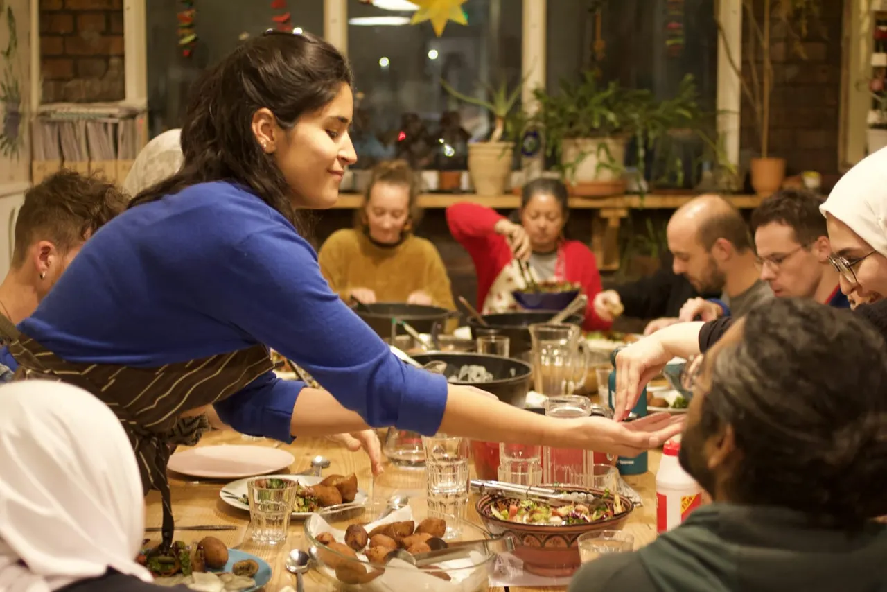 A woman in a blue top serves food to a group of people seated around a wooden dining table during a cooking class or communal meal.