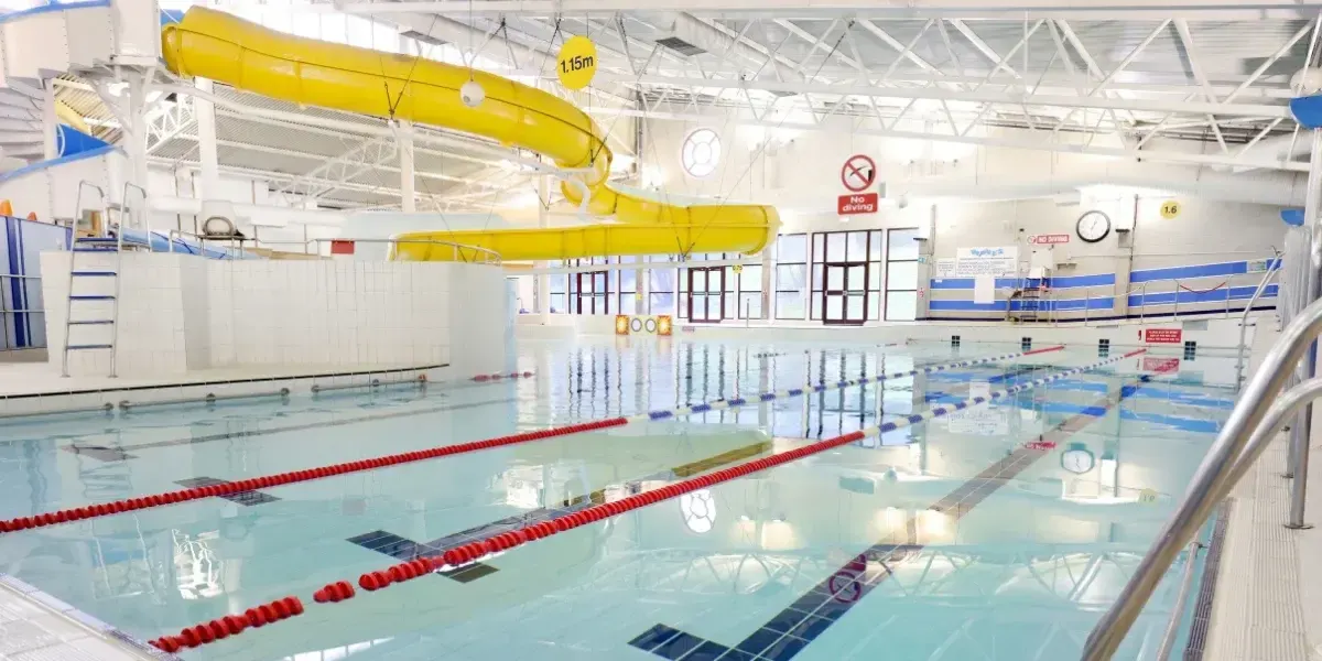 Indoor swimming pool with multiple lanes divided by red lane ropes, yellow water slide overhead, and white partition walls.