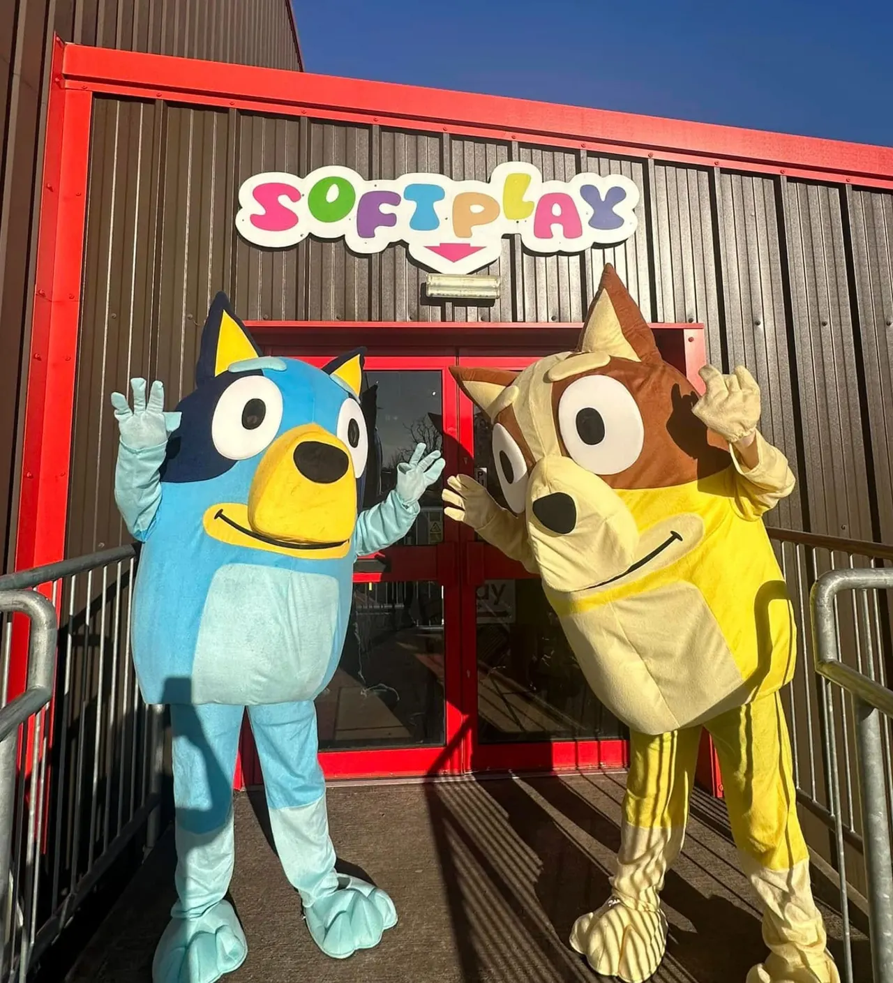 Two colourful cartoon character mascots in costumes stand outside a soft play venue entrance on a sunny day.