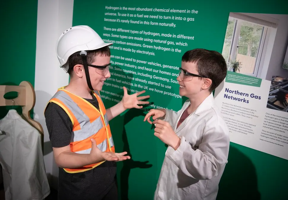 A young person wearing a hard hat and high-visibility vest explains hydrogen energy to a visitor in front of a green educational panel.