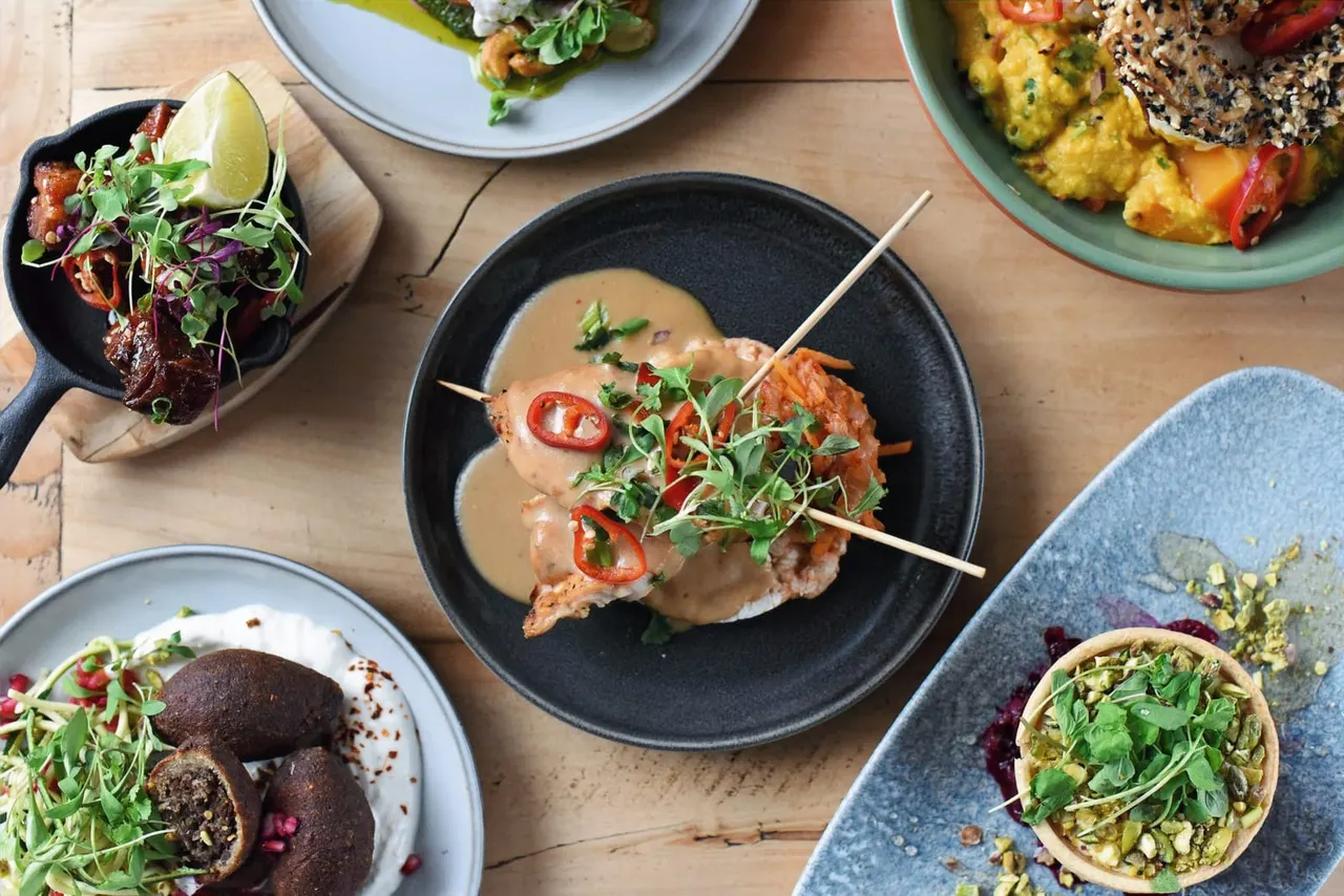 Overhead view of restaurant table with multiple plated dishes including skewered meat in sauce, chocolate desserts, and vegetable-based dishes.