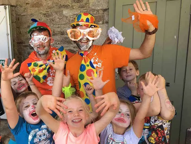 Two adults dressed as clowns with face paint and colourful costumes pose with a group of smiling children, all with their hands raised in celebration.