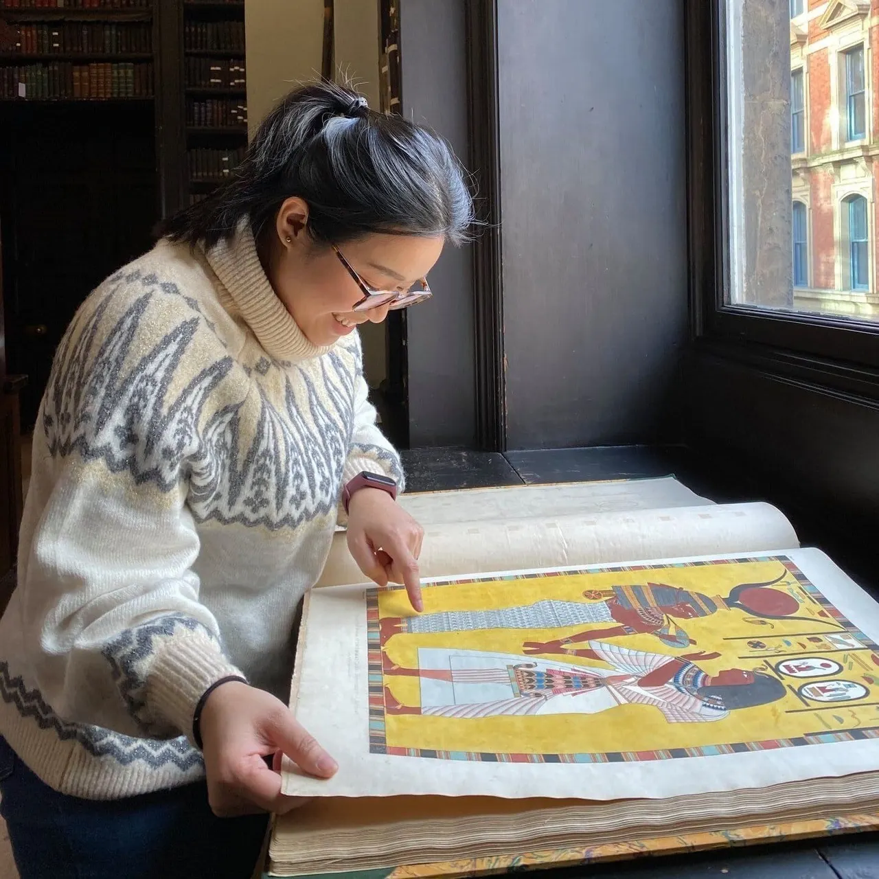 Woman examining an open illustrated manuscript or art book in a historic library interior with bookshelves and windows.
