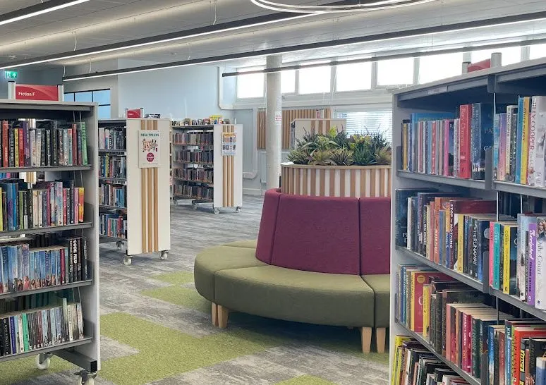Interior of a library with bookshelves, a curved two-tone sofa and potted plants in a modern, bright space.