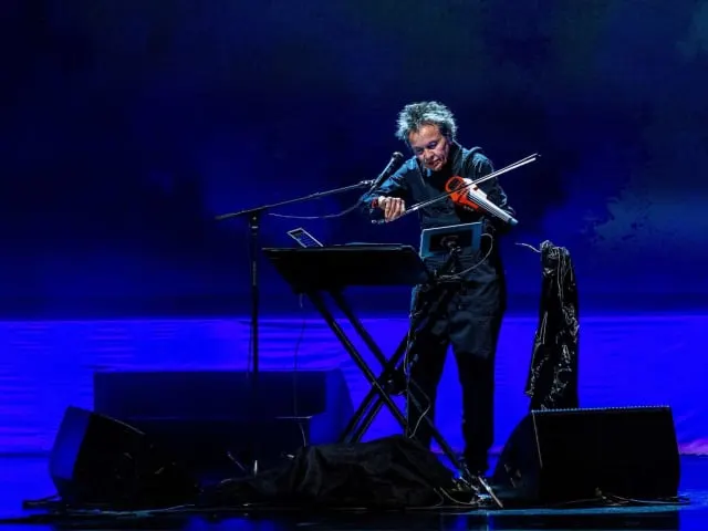 Musician performing at keyboard on stage with blue lighting, playing violin-like instrument simultaneously.