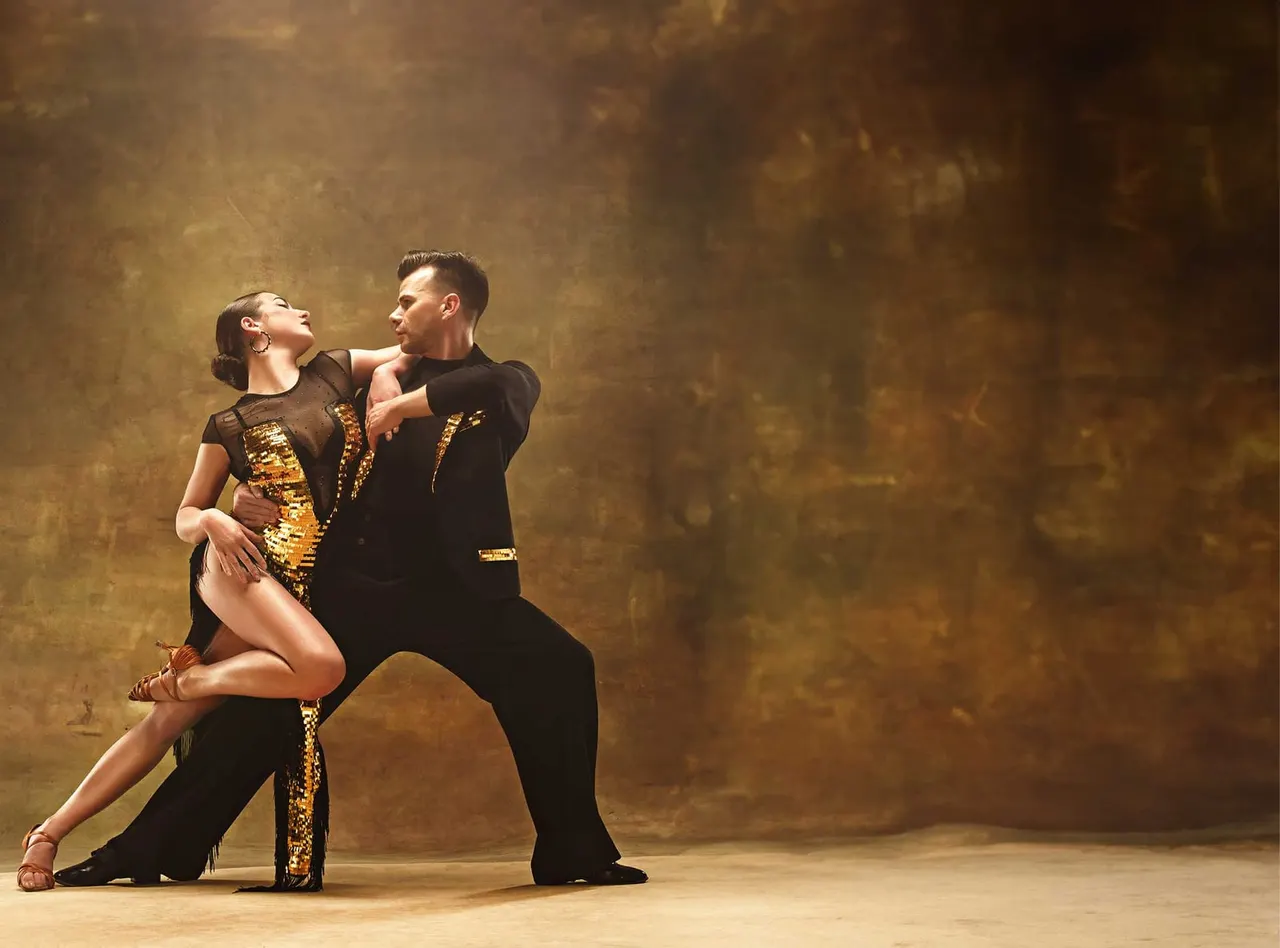 Two dancers performing Latin dance, the woman in a black and gold sequined dress and the man in black with gold trim, against a textured golden-brown backdrop.
