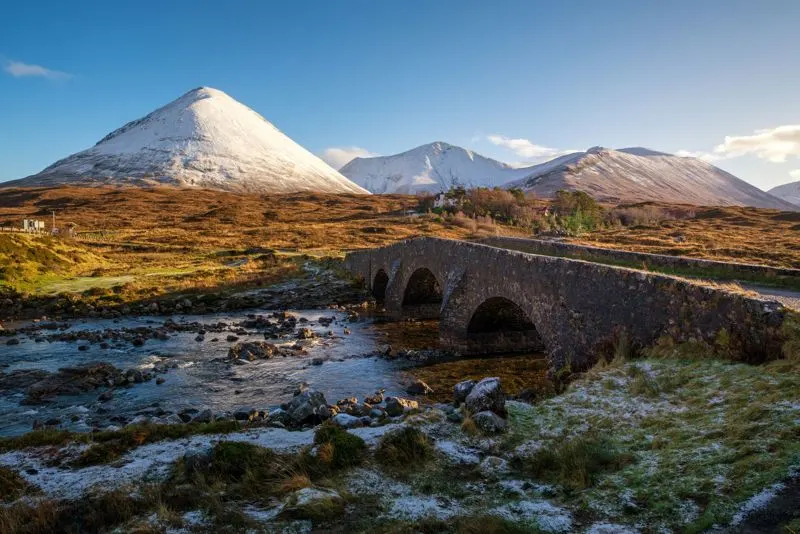 Stone bridge spanning a stream with snow-capped mountains in the background under clear blue sky.