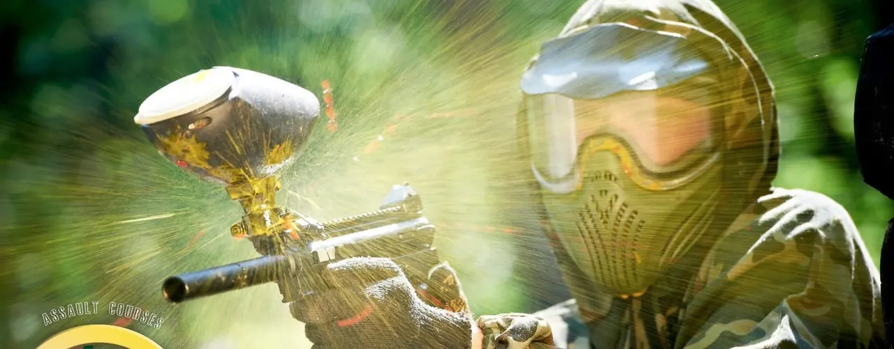 A person in protective goggles and mask aims a paintball marker during an outdoor assault course activity.