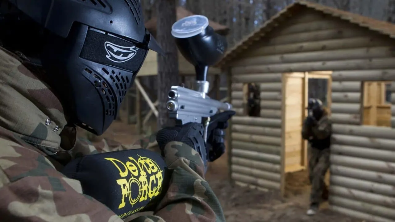 Player in paintball gear holding marker gun with protective mask, aiming at wooden buildings during game.