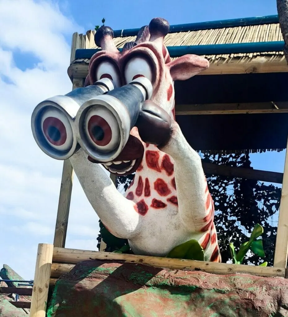 Oversized novelty giraffe statue with binoculars holds two visitors aloft against blue sky.