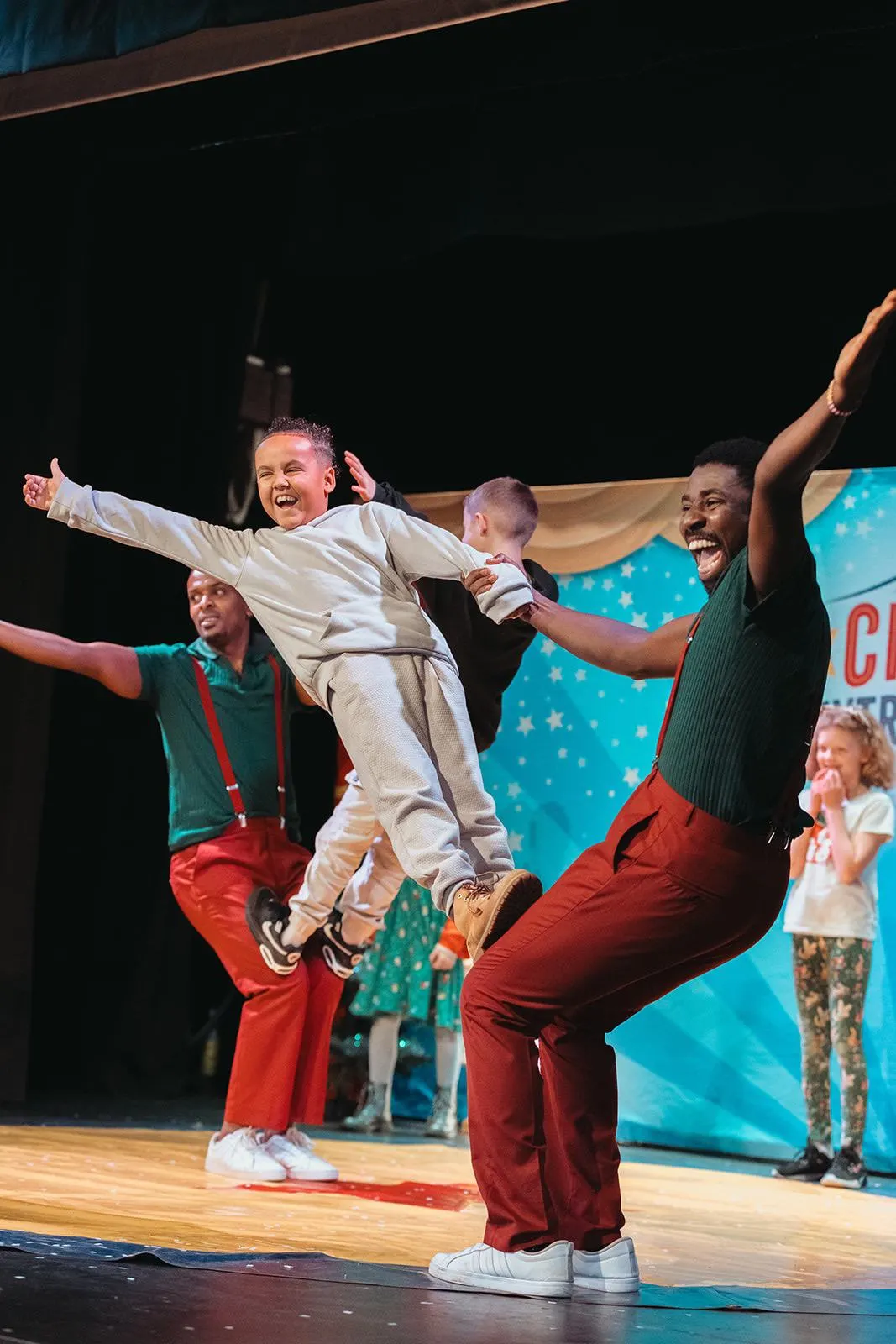 A young child in cream-coloured clothing is lifted high by an adult performer on stage, arms outstretched joyfully, surrounded by other smiling performers during a circus show.