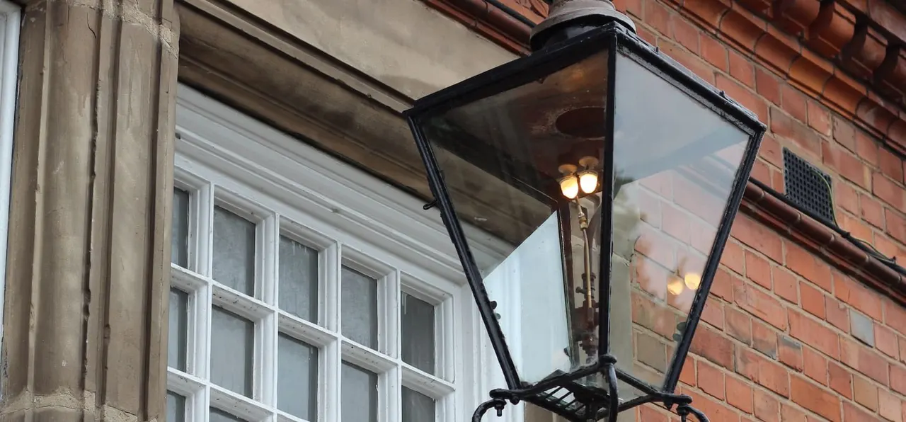 A traditional black metal lantern with glowing lights mounted on a brick building exterior beside a multi-paned window.