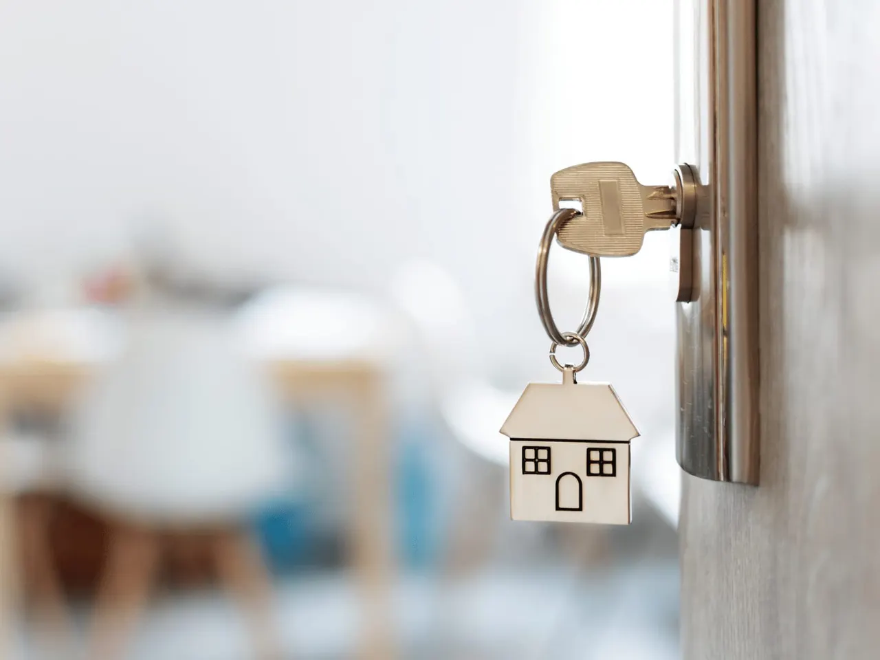 House-shaped keyring hanging from a key in a door lock, with a blurred interior space in the background.