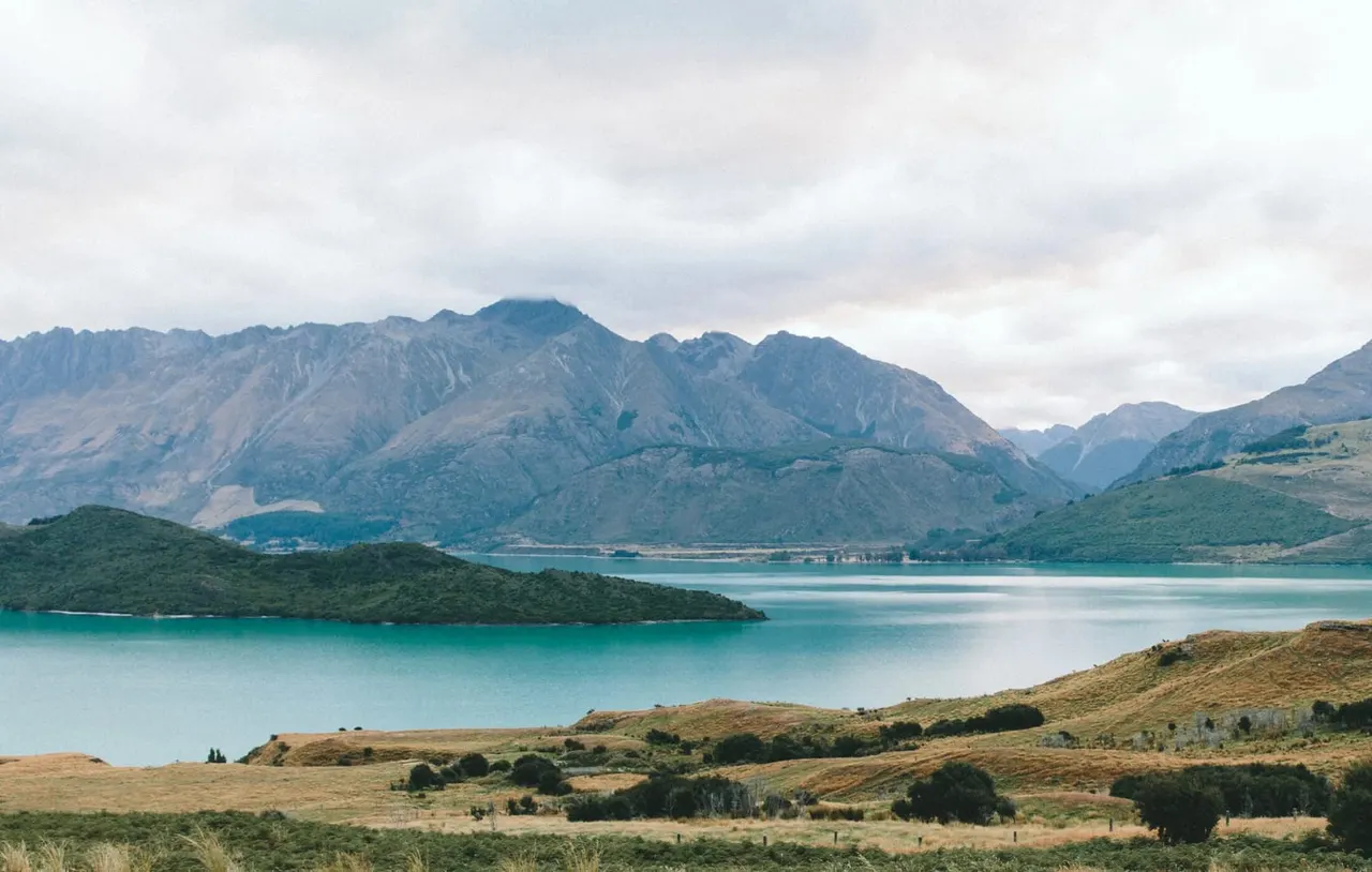 Turquoise lake surrounded by forested hills and snow-capped mountains under overcast sky.