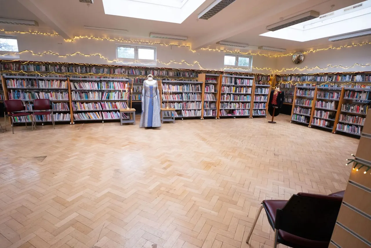 Library interior with wooden bookshelves lining walls, fairy lights strung overhead, and a mannequin wearing a blue gown displayed centrally.