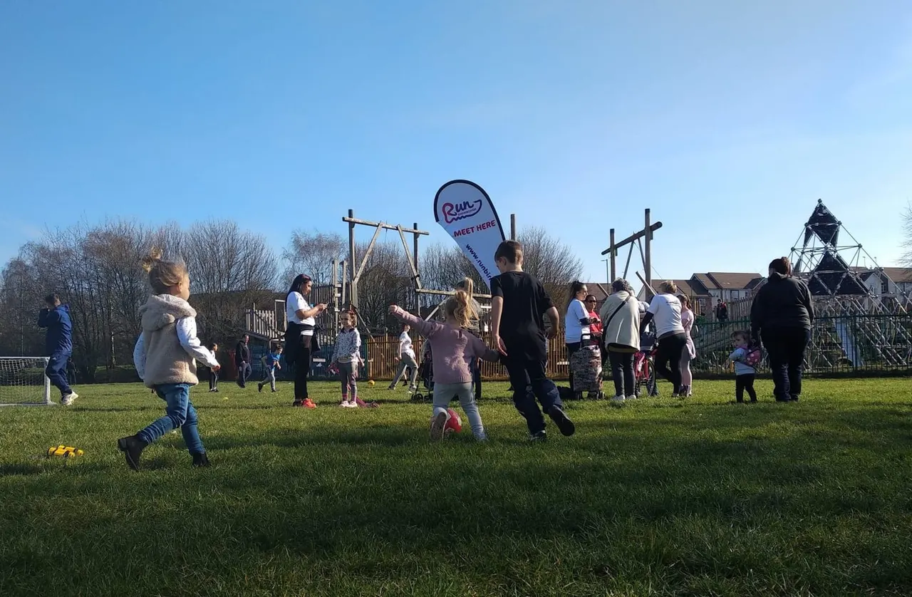 Children running and playing in a grassy park with wooden playground equipment and a "Meet Here" banner on a clear day.