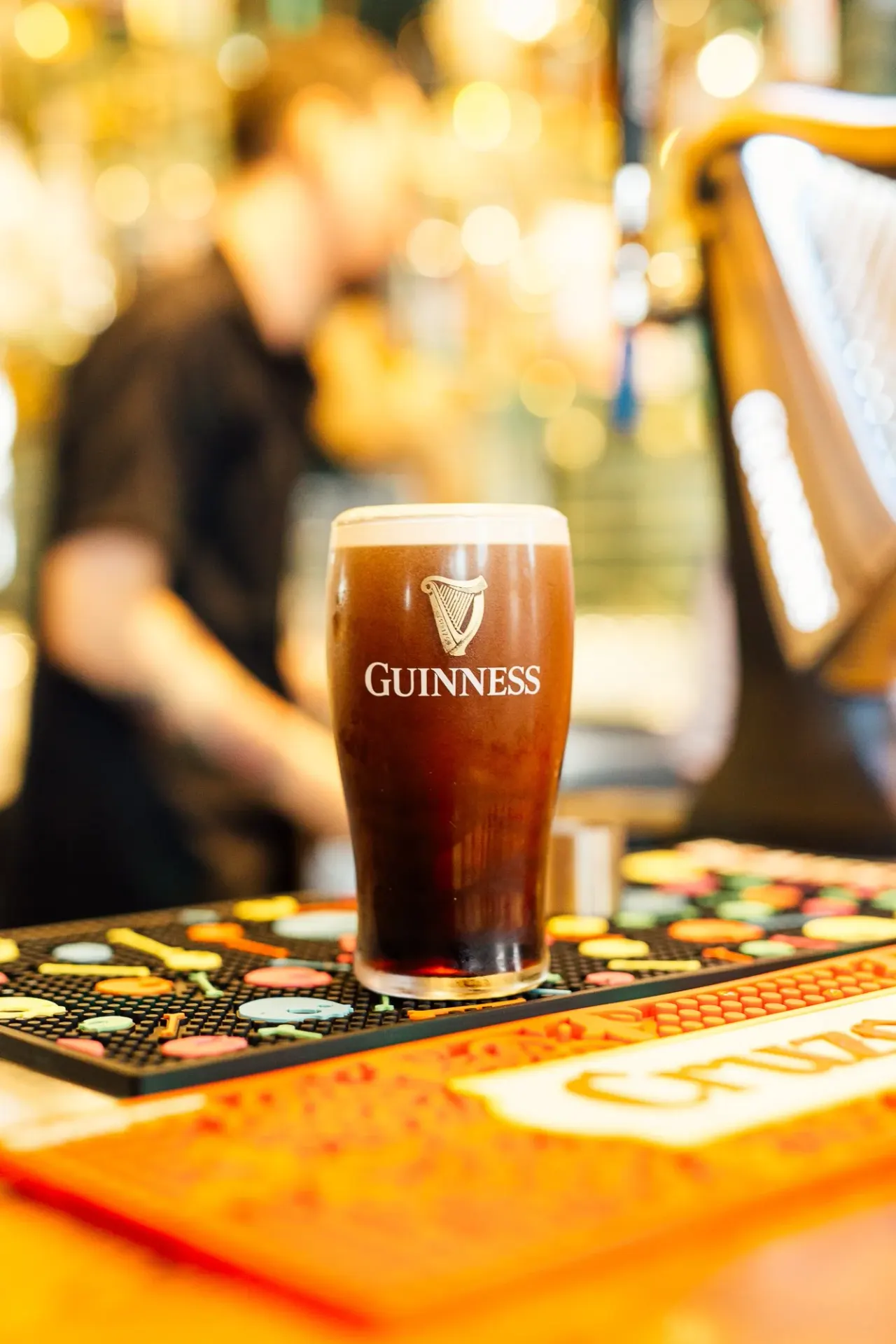Pint of Guinness on a bar table with blurred patrons and warm lighting in the background.
