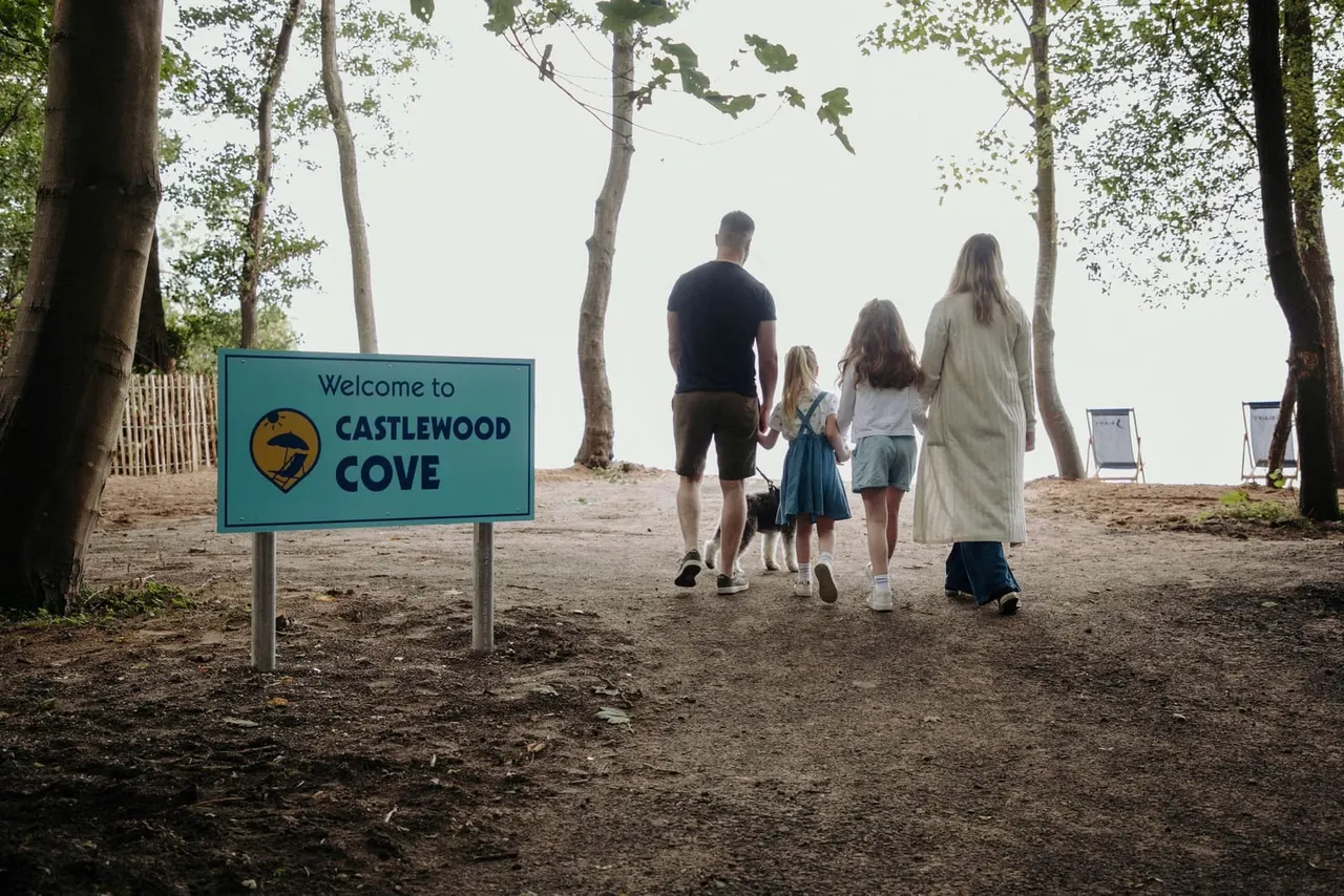Family walking along a woodland path past the Castlewood Cove welcome sign towards a beach area.
