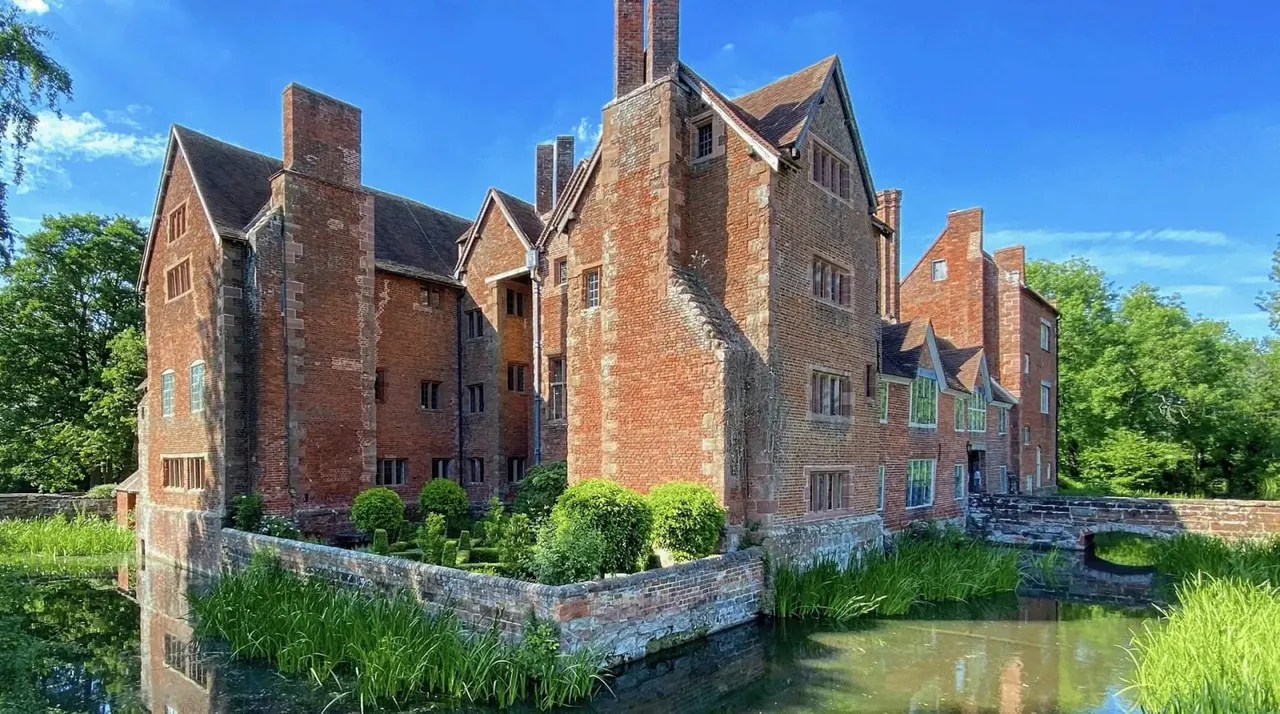 Historic red-brick manor house with multiple chimneys surrounded by moat and green grounds under blue sky.
