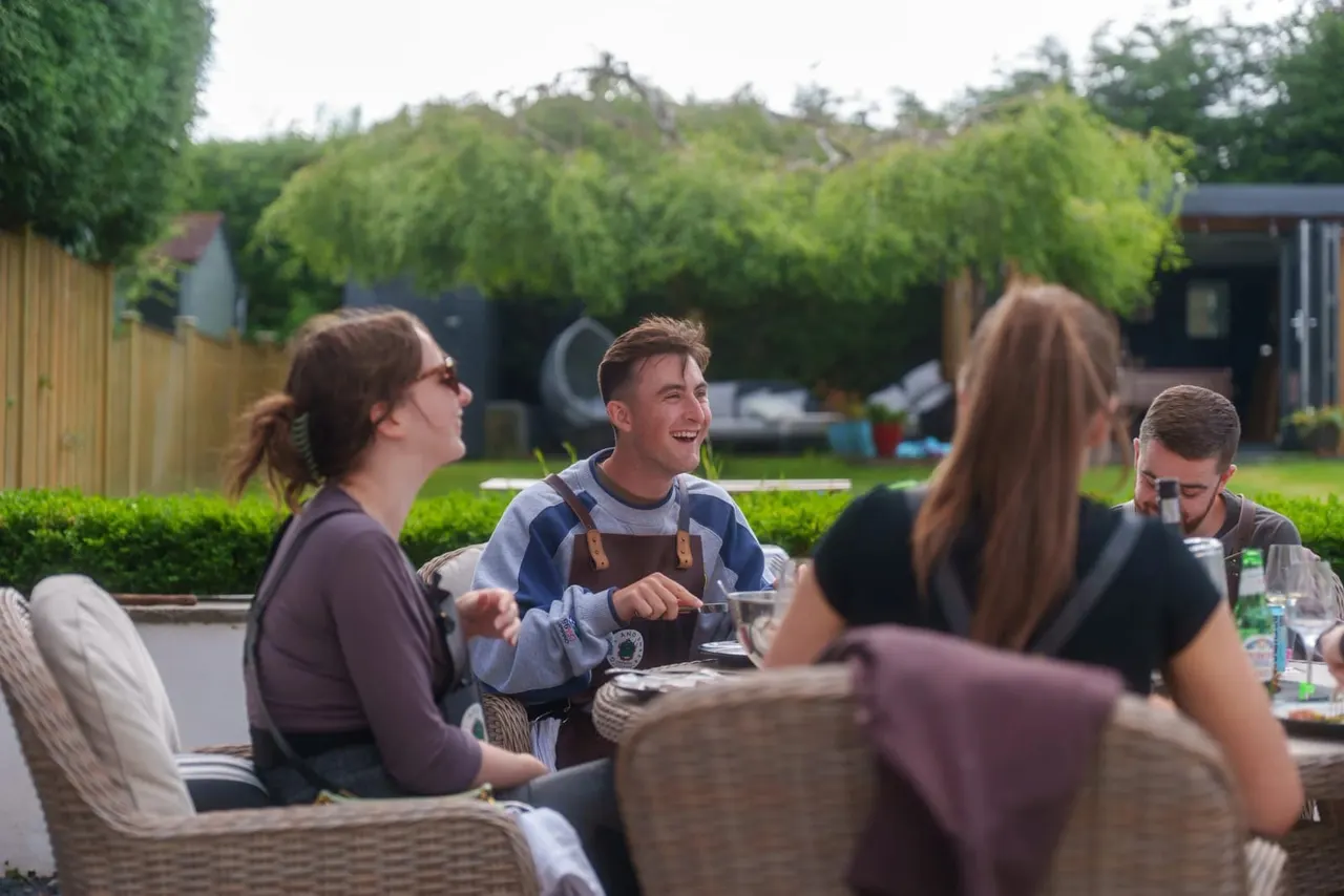 Group of adults enjoying a meal and conversation at outdoor seating in a garden setting with hedges and outbuildings.