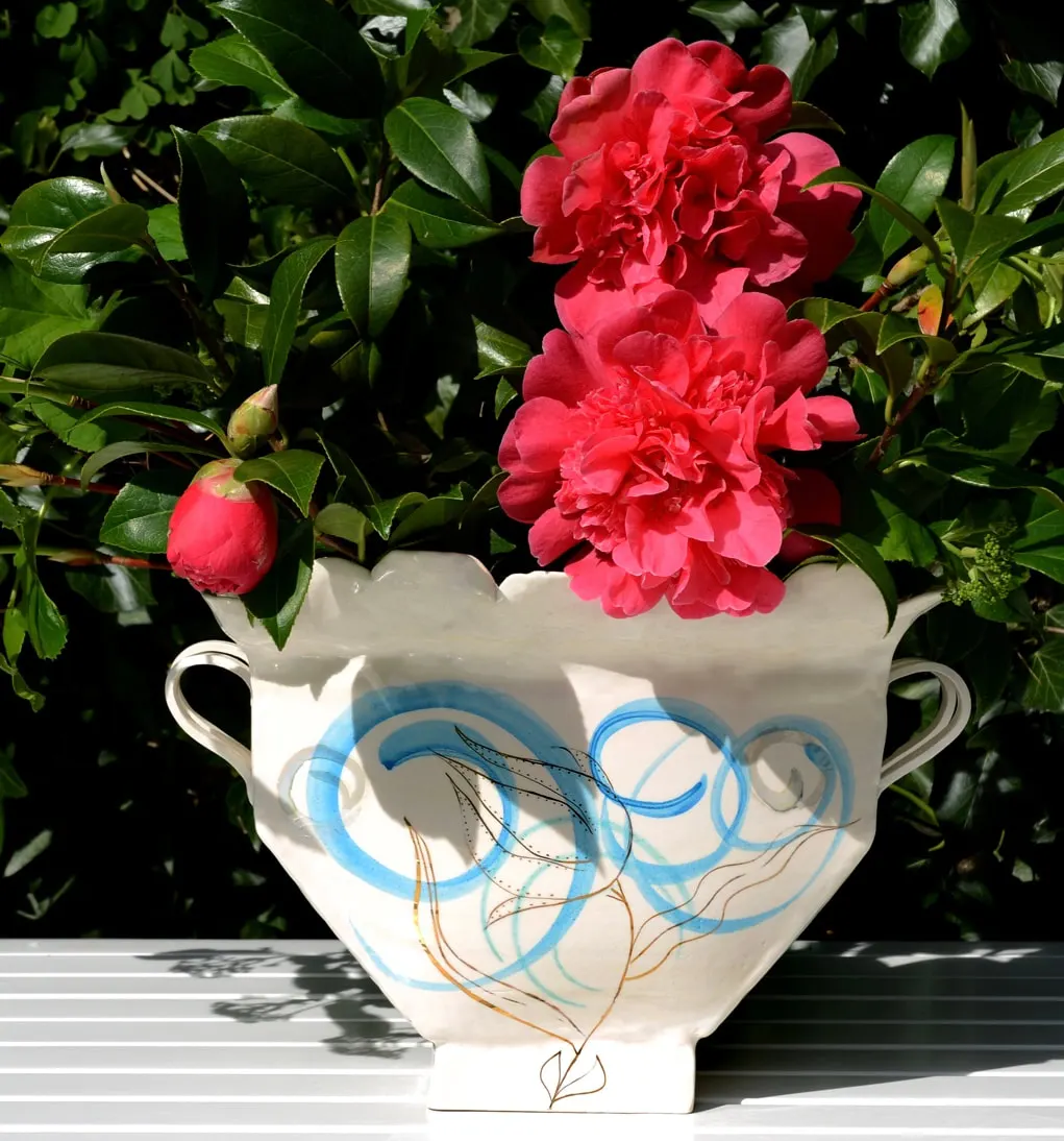 Bright red peonies arranged in a white ceramic vase with blue decorative patterns, displayed outdoors against green foliage.