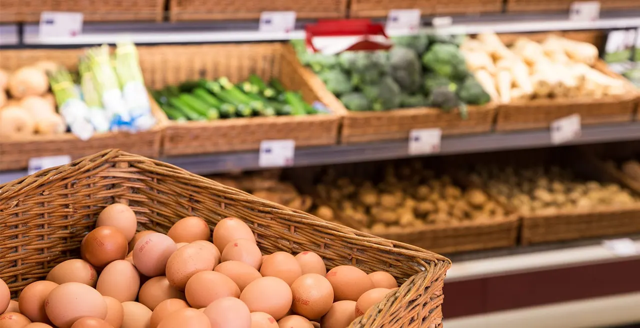 Wicker basket of brown eggs displayed in foreground with farm shop produce shelves of vegetables blurred in background.