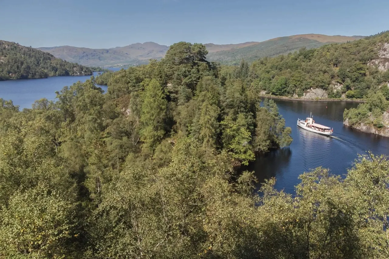 A passenger steamer navigates a loch surrounded by dense woodland and mountains under clear blue sky.
