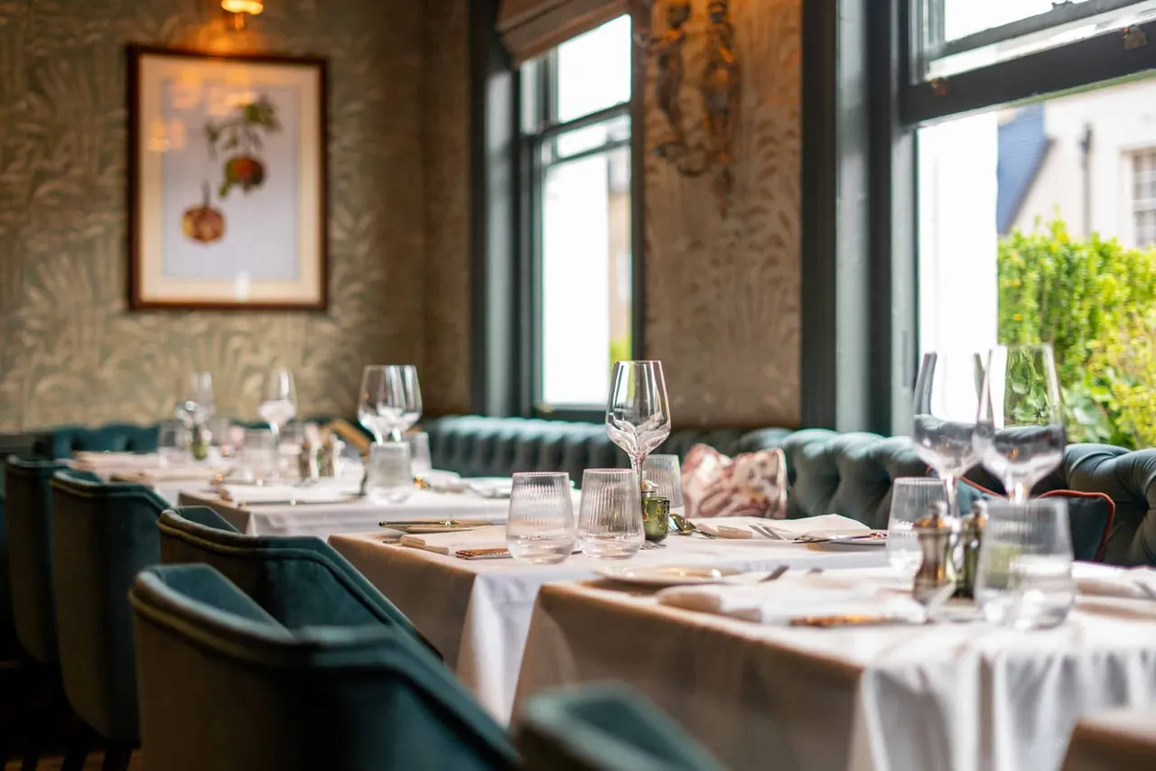 Elegant restaurant dining room with set tables, wine glasses, and teal banquette seating beside large windows.