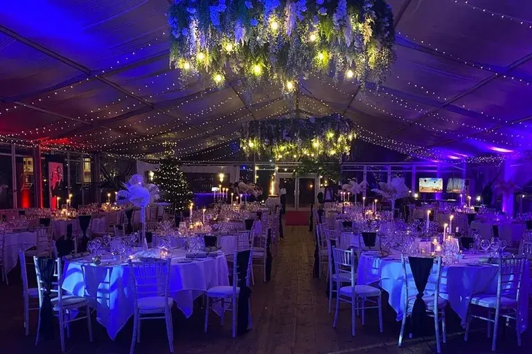 Elegant banquet hall decorated with purple and blue lighting, white tablecloths, hanging floral installations, and string lights for an event.