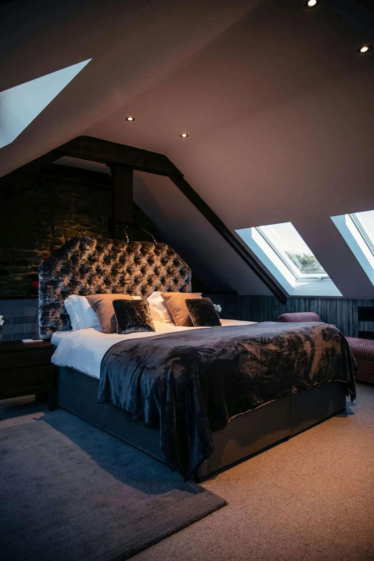 Modern attic bedroom with double bed, patterned headboard, skylight windows and exposed beams in warm lighting.