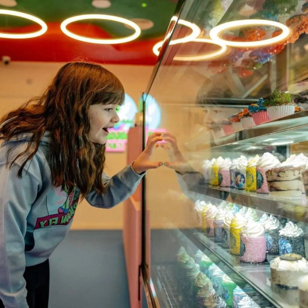 Child looking at cakes and desserts displayed in a refrigerated case beneath neon signage.