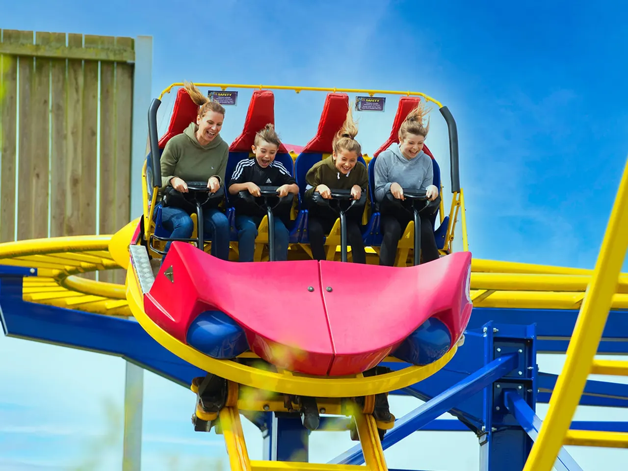 Four children on a red and yellow roller coaster ride with their hands up, smiling against a blue sky.