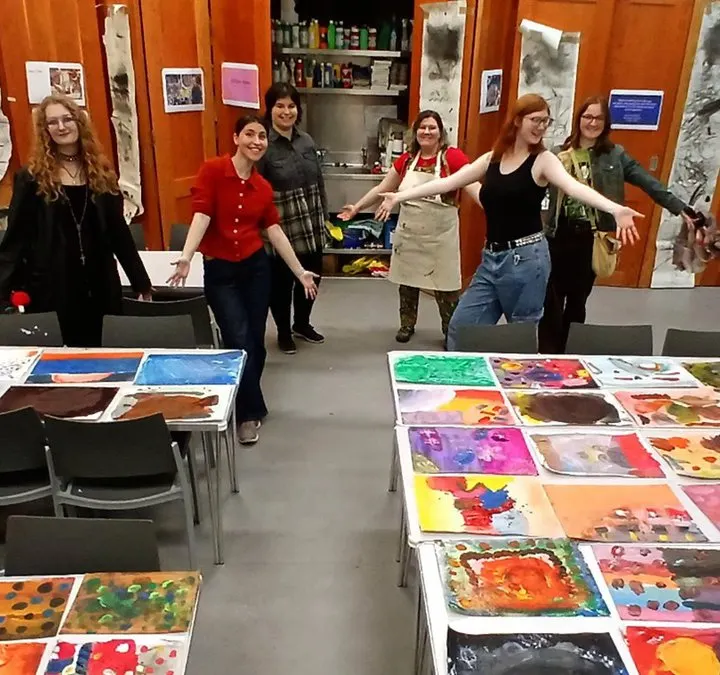 Group of six young people posing in an art studio with colourful paintings displayed on tables and bright orange walls.