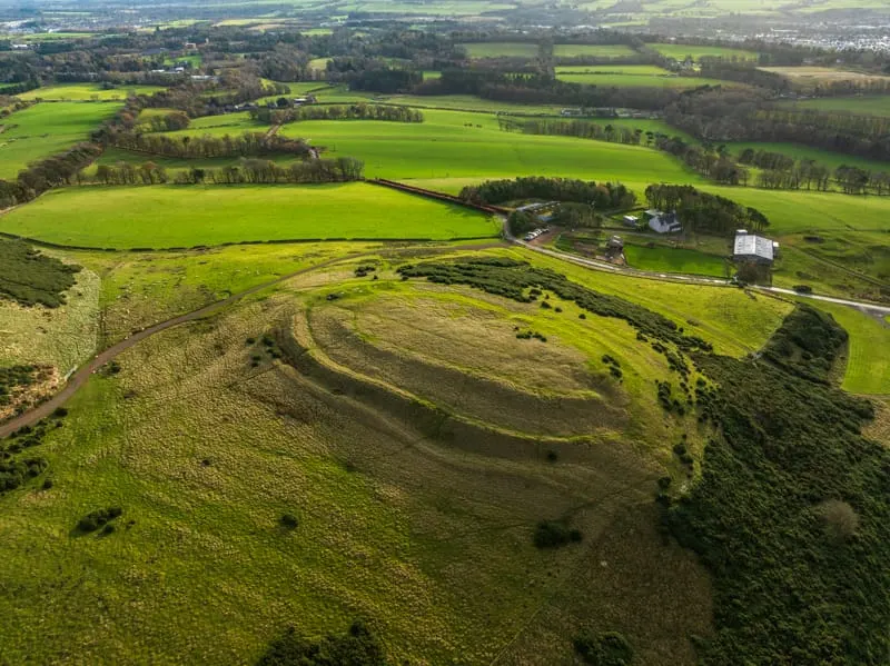 Aerial view of an ancient hill fort with concentric ramparts surrounded by green fields and woodland.