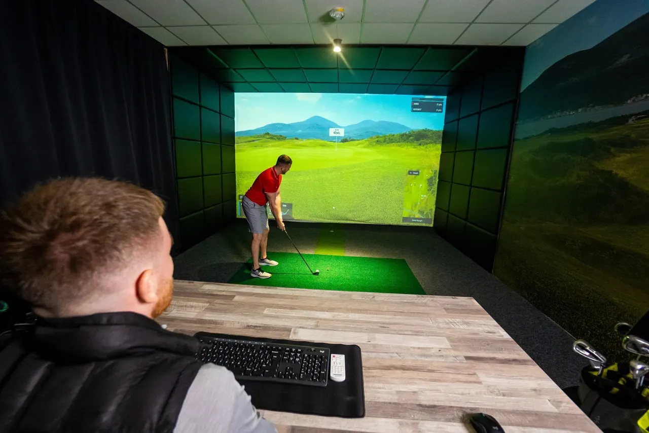 Two people use an indoor golf simulator with a large screen displaying a mountainous landscape and putting green.