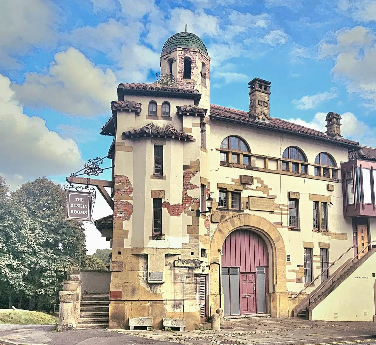 Historic sandstone building with domed tower and "The Ruskin Rooms" sign, set against blue sky with white clouds.