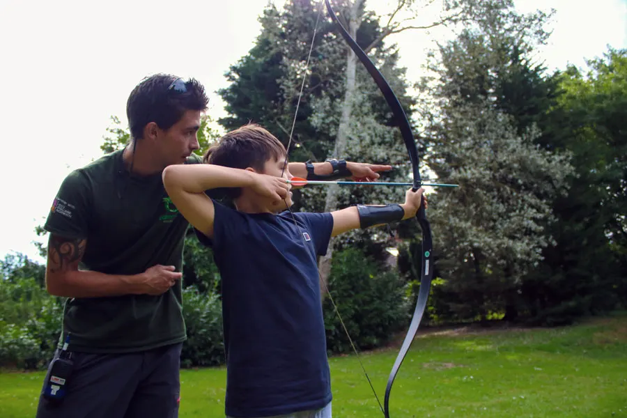 Instructor supervising a young person aiming a bow and arrow in a grassy outdoor setting surrounded by trees.