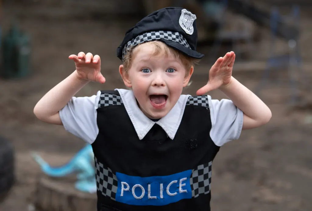 Young child in police officer fancy dress costume with arms raised, smiling with mouth open.