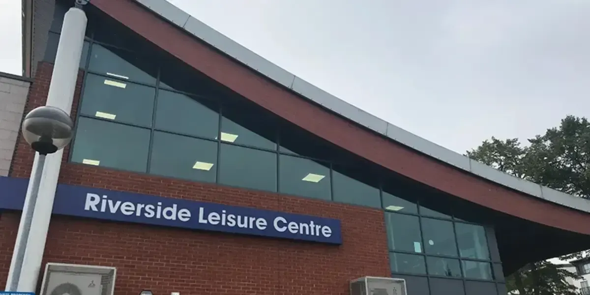Exterior of a modern leisure centre building with red brick and large windows, signage reading "Riverside Leisure Centre" visible on the facade.