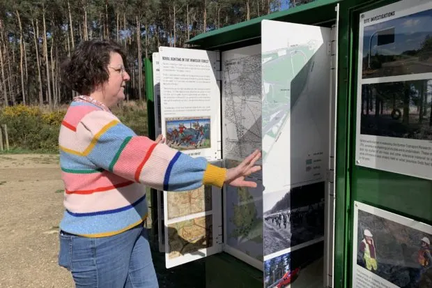 Person in colourful striped jumper reading an outdoor information board in a forest clearing.