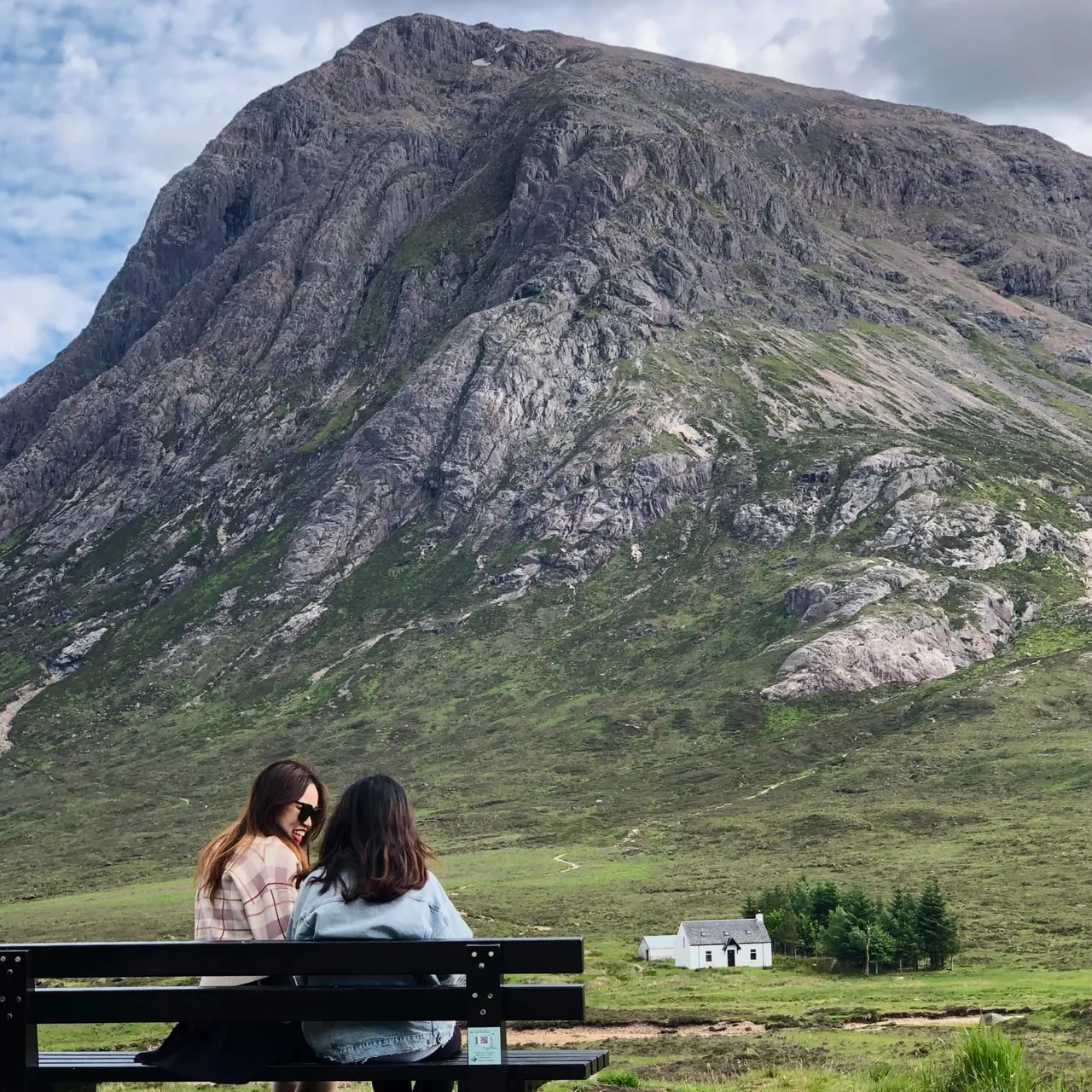 Two people sitting on a bench facing a dramatic rocky mountain with green slopes and a white cottage in a Highland valley.