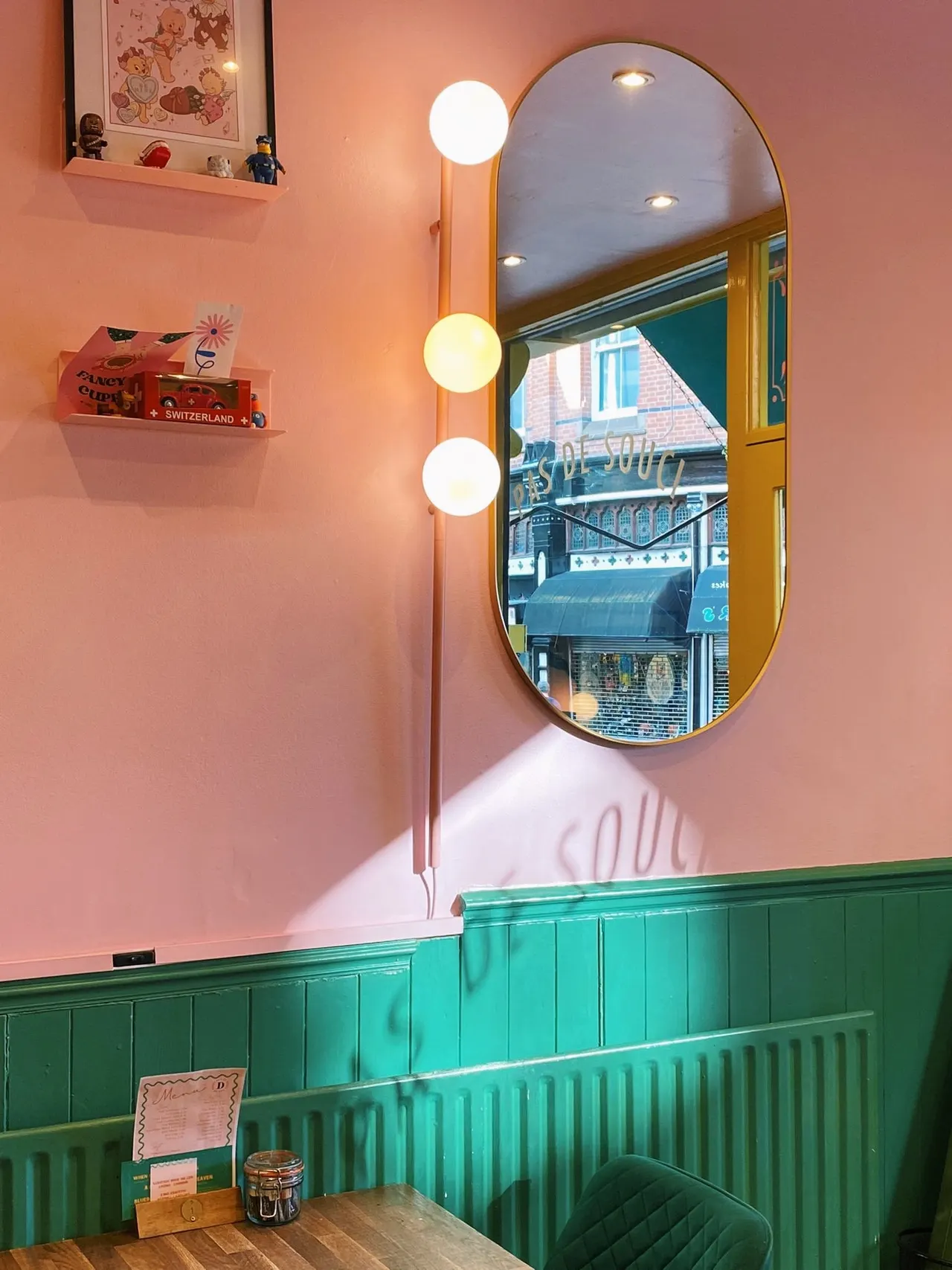 Pink and green interior with illuminated mirror, globe wall sconces, shelving with collectibles, and street view through arched window.