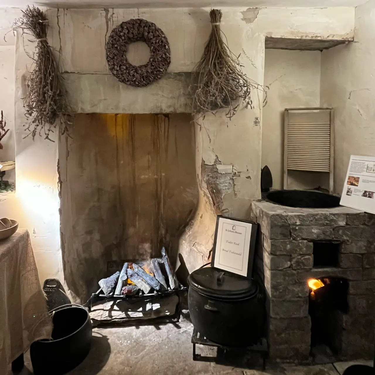 Historic interior display showing traditional hearth with black cooking pots, dried herbs and wreath decorations on whitewashed walls.