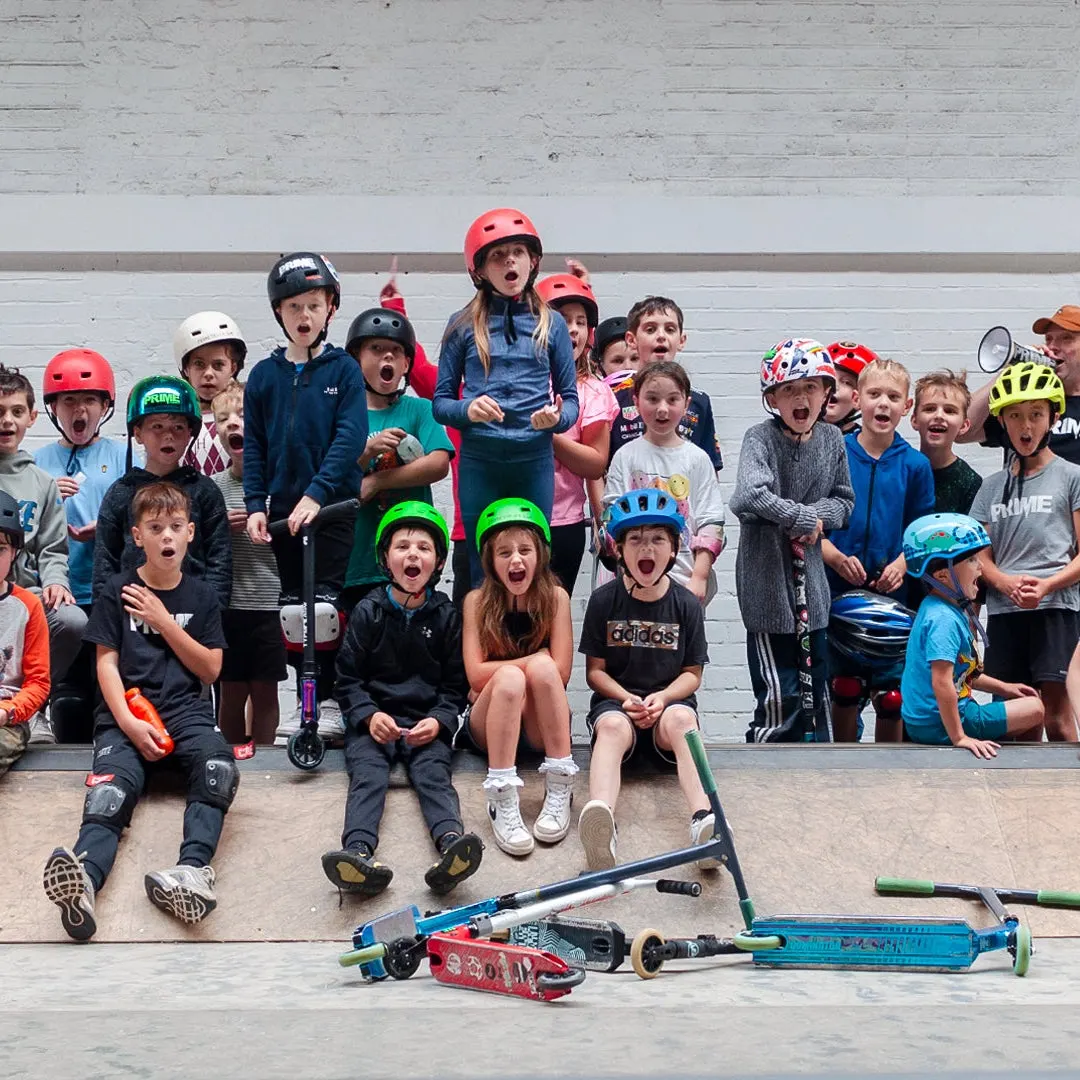 Large group of children wearing safety helmets gathered at a skate park, many cheering and posing with scooters and skateboards.