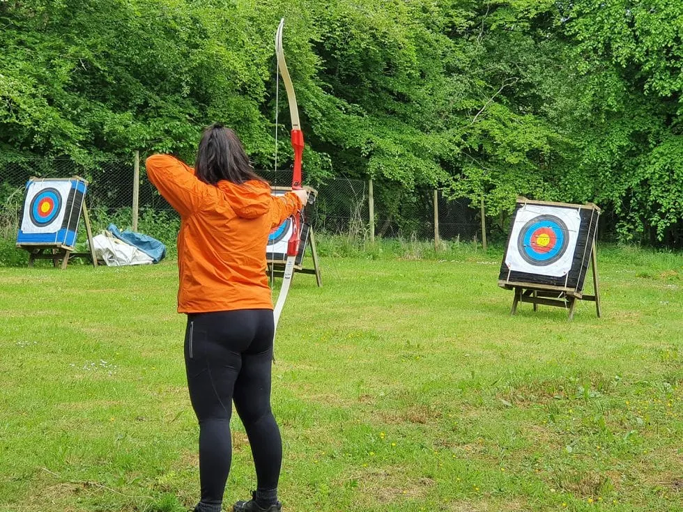 Person in orange jacket drawing a bow and arrow at targets on a grassy field surrounded by trees.