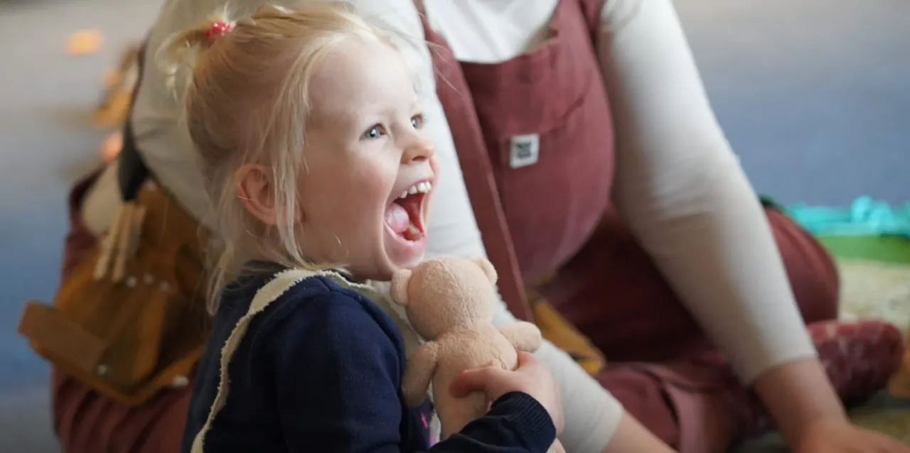 Young child holding a teddy bear, smiling with delight whilst being held by an adult carer.