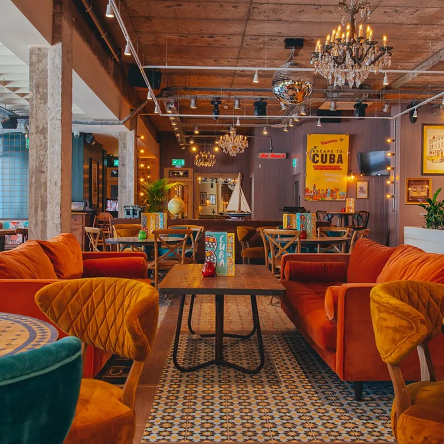 Colourful interior of a leisure venue with orange velvet seating, wooden tables, chandeliers, and Cuban-themed décor on the walls.