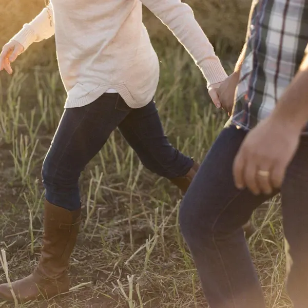 Two people in boots and casual clothing walking through a grassy field at dusk.